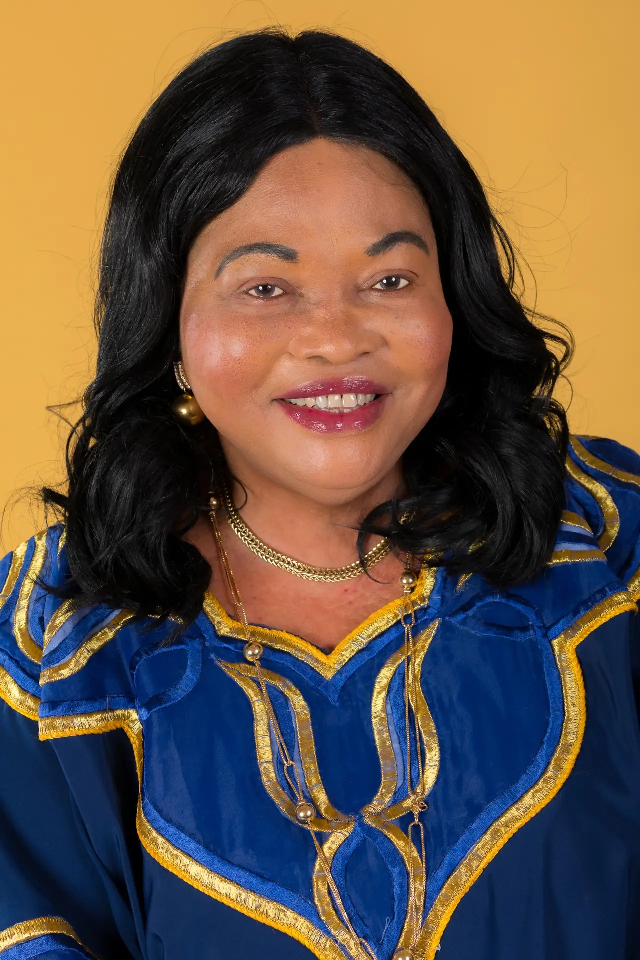 Portrait of a smiling woman with long black hair in a blue and gold embroidered outfit, yellow background.