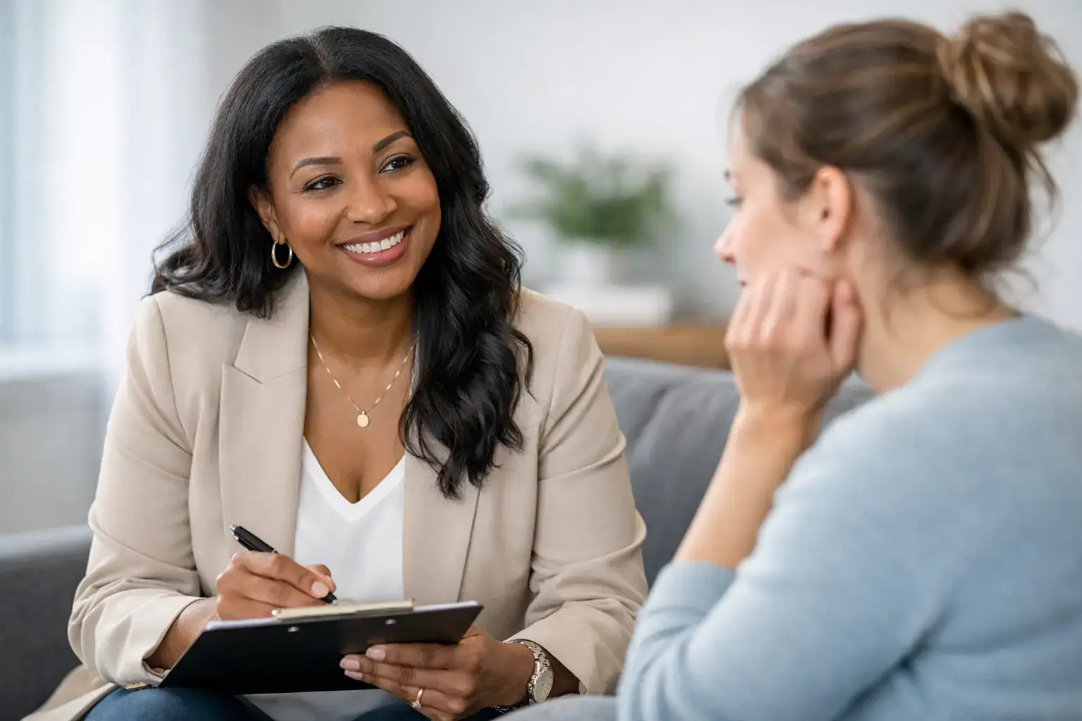 Two women on a sofa in a bright room; one with a clipboard smiles as she talks to the other client.