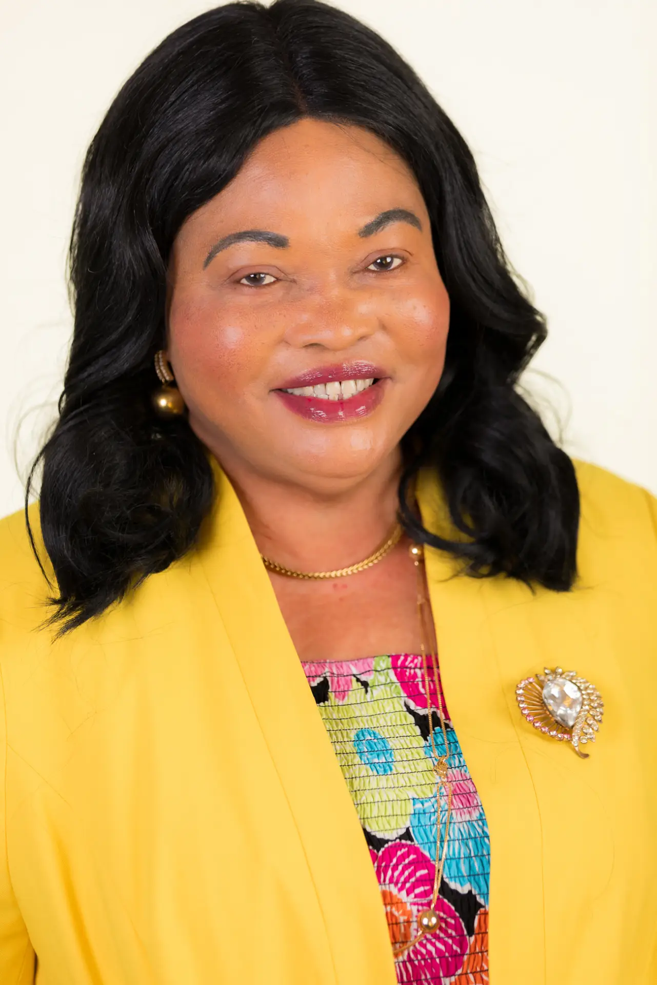 Portrait of a smiling woman with long black hair, wearing a bright yellow blazer and a colorful patterned top, gold jewelry visible behind her shoulders.