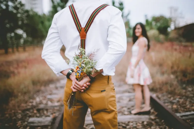 Man in white shirt and mustard suspenders holding a small bouquet behind his back on train tracks, woman in a floral dress blurred in the background.